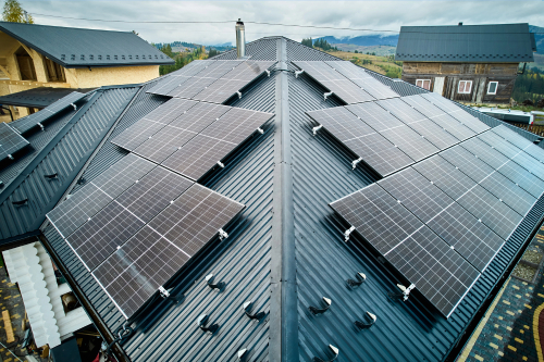 A large, black metal roof covered in rows of solar panels on a modern house in a hilly, rural area with other homes and trees in the background under a cloudy sky.