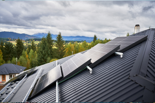 Solar panels mounted on a dark metal roof with a scenic backdrop of pine trees, rolling hills, and distant mountains under a cloudy sky.
