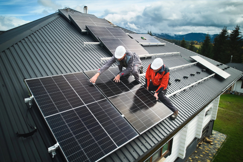 Two technicians in safety helmets secure large solar panels on a sloped black metal roof, carefully aligning the setup.