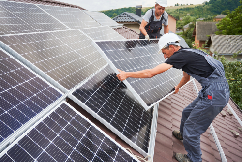 Two men collaborating on a rooftop, installing solar panels in rooftop with tools in hand and safety measures in place.