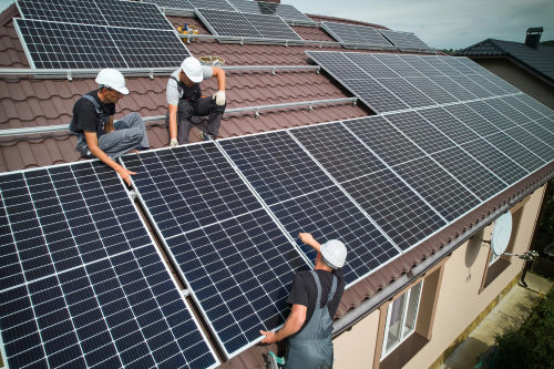 Three men collaborating on a rooftop, installing solar panels with tools and equipment around them.