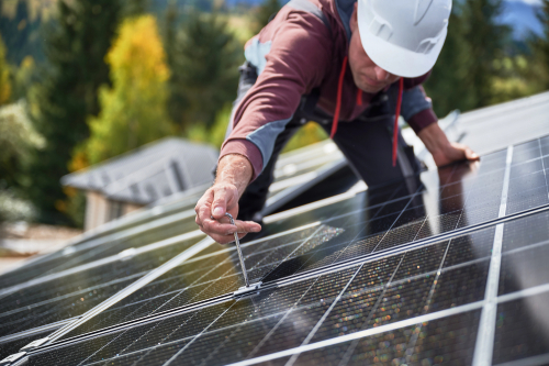 A worker wearing a hard hat and helmet is engaged in the installation of a solar panel on a rooftop.