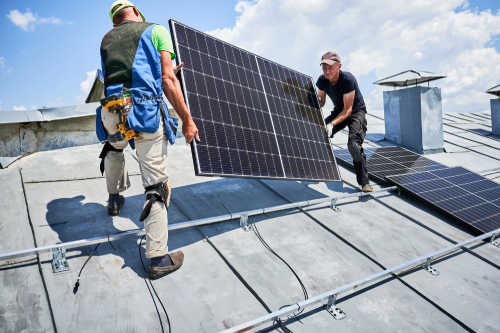 Two men collaborating on a rooftop, installing solar panels with tools in hand and safety measures in place.